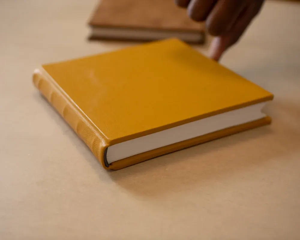 Brown hardcover book with cream pages on a table, with stacked books in the background.